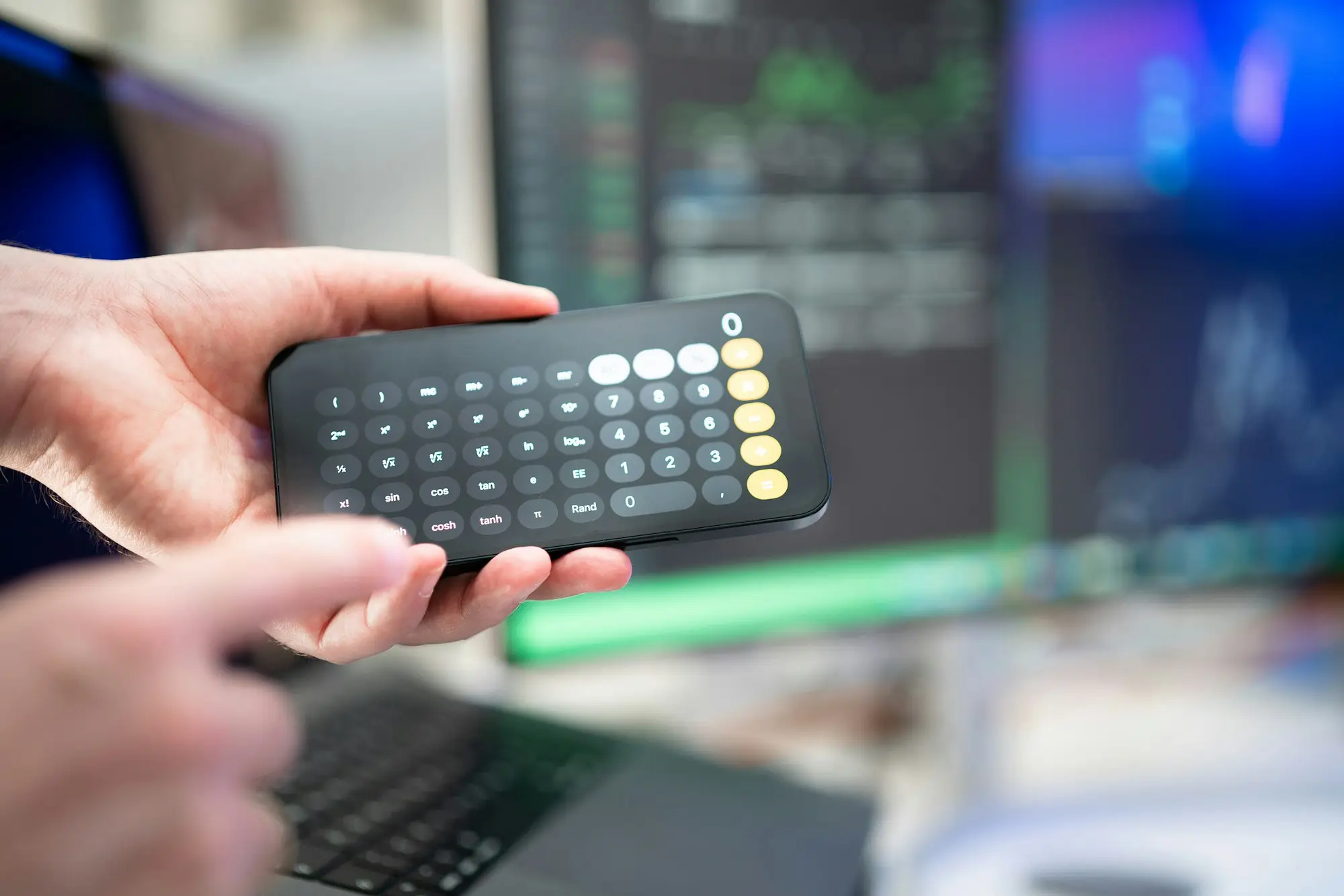 Hands holding a calculator in front of a laptop and monitor while adding up business debts