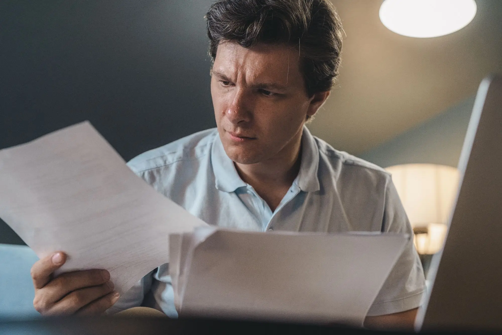Business owner frowning as he reads through a printed court claim at his desk, with a second set of papers beside his laptop