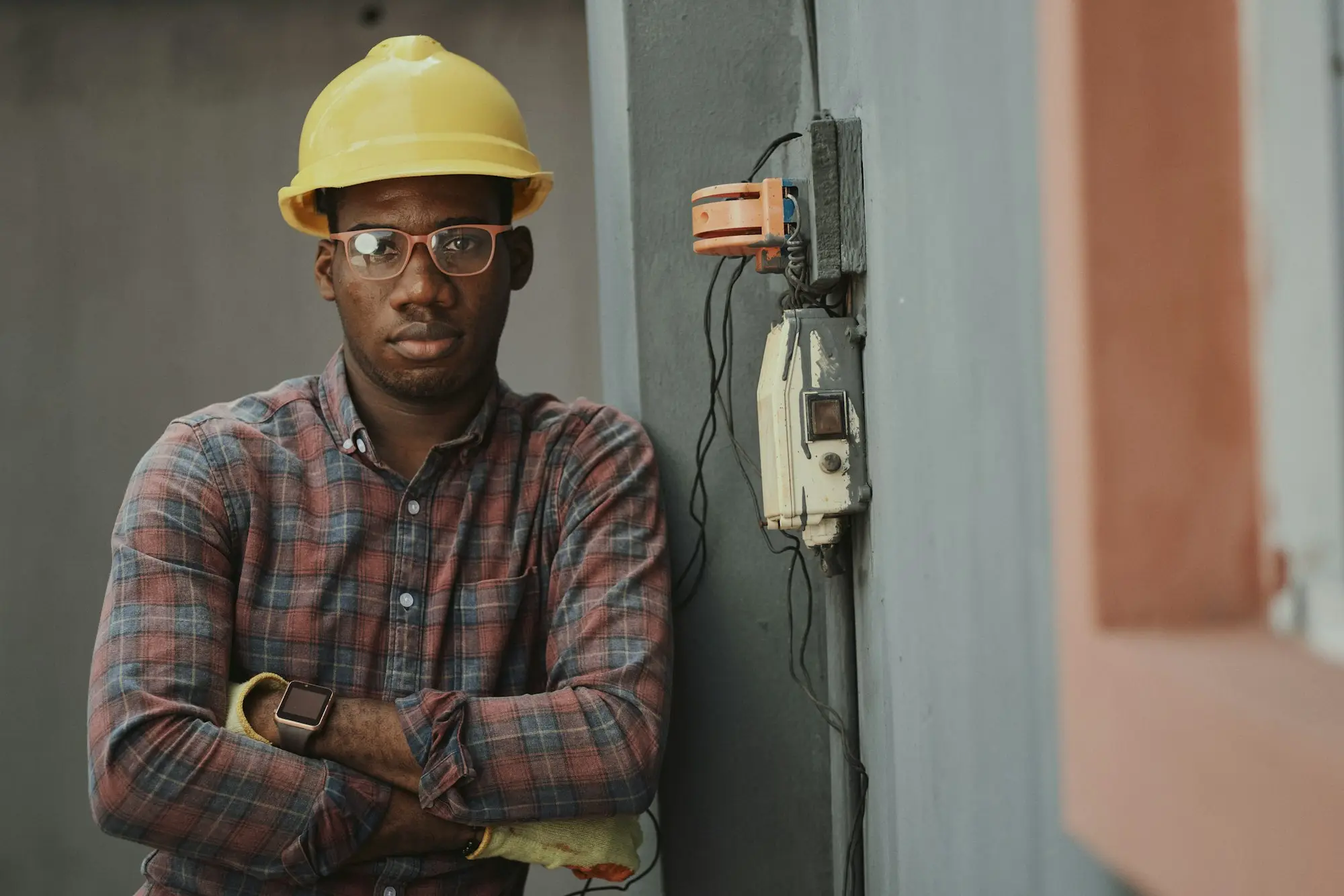 Electrical contractor in a hard hat and flannelette shirt standing with arms folded beside a switchboard, weighing up a supplier's demand under a personal guarantee